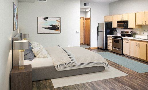1 BR Apartments in Downtown Sacramento CA - Fremont Mews Apartments - Studio Bedroom Area with Wood-Style Flooring, Area Rugs, Bed, Nightstands with Lamps, Wall Art, and View of Kitchen at FREMONT MEW