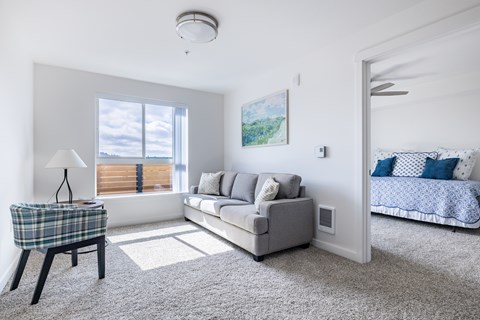 A well-lit, modern living room with a grey sofa, a small table with a lamp, and a bed with a blue and white comforter in the background.