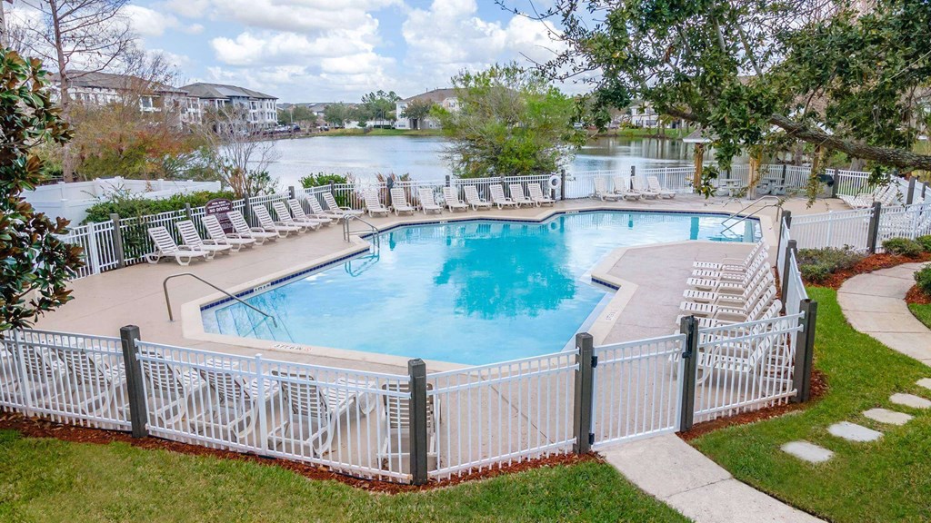 A pool surrounded by a white fence with a view of a lake and houses in the background at The Villages on Millenia Apartments, Orlando, Florida