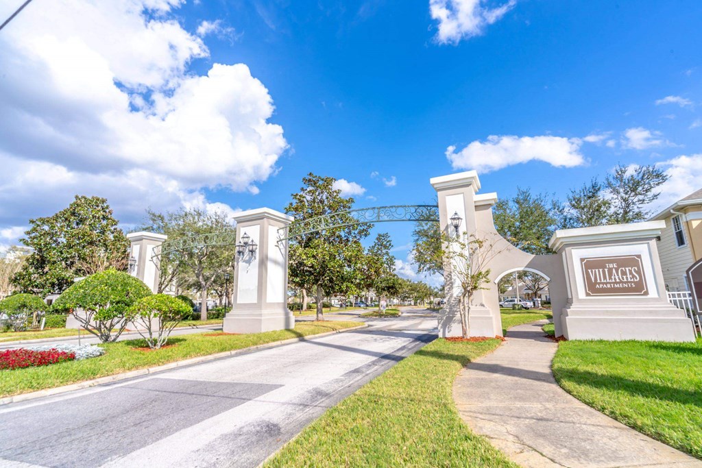The entrance to a place called Villages is shown with a clear blue sky above at The Villages on Millenia Apartments, Orlando, FL