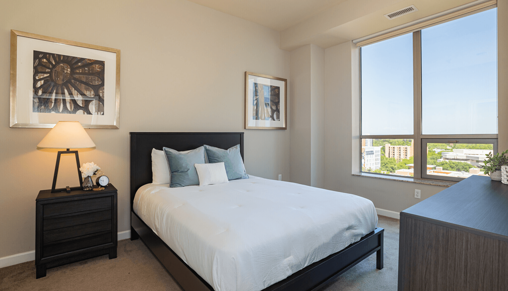 Sacramento, CA Luxury Apartments - Spacious Bedroom With Carpet Flooring, Bed, Nightstand with Lamp, Dresser, High Ceilings, And A Large Window