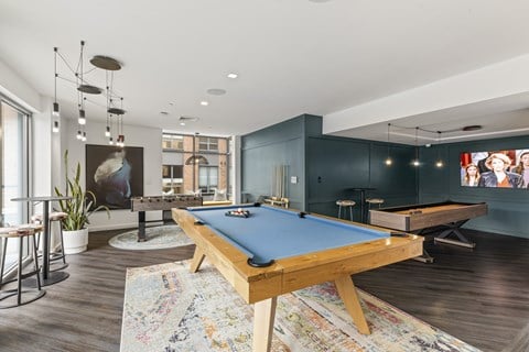 A pool table is in the middle of a room with a bar stool and a potted plant. at Trio Newton Apartments, Massachusetts