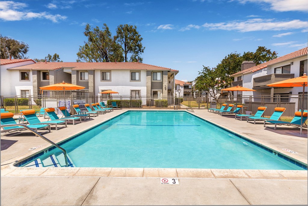 Pet-Friendly Apartments in Moreno Valley, CA - Sorelle Apartments - Classic Rectangular Swimming Pool Flanked by Rows of Blue Lounge Chairs and Orange Umbrellas with Views of the Apartment Homes