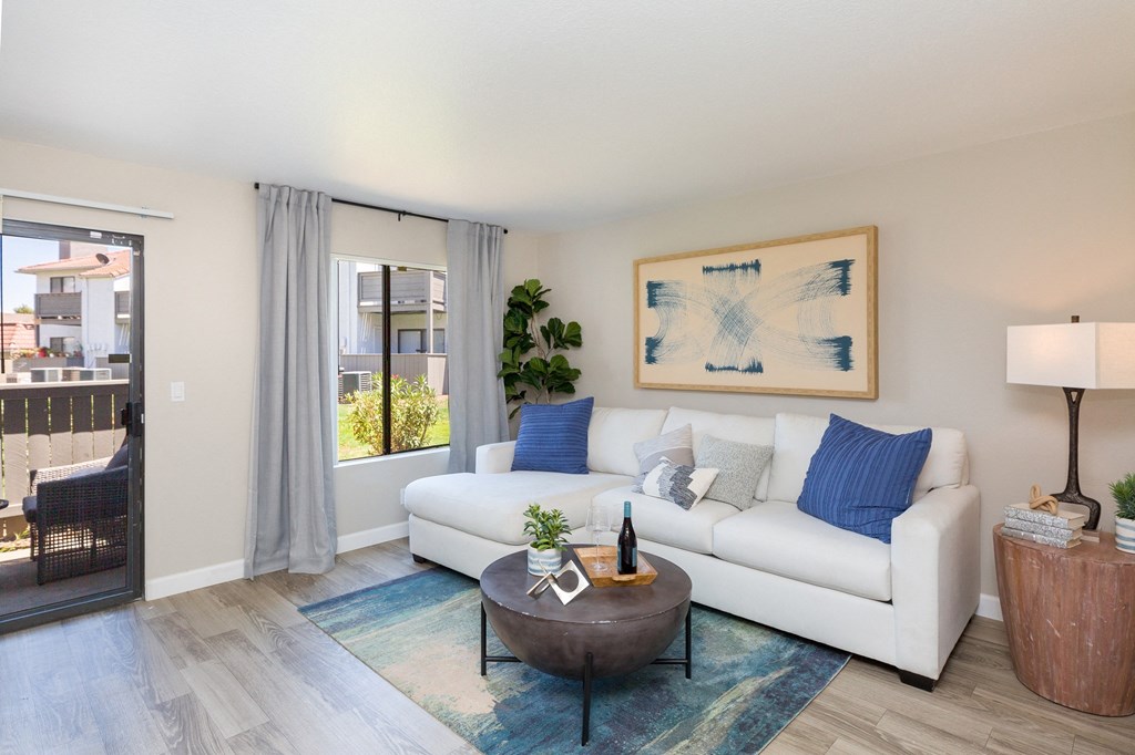 Dog-Friendly Apartments in Moreno Valley, CA - Sorelle Apartments - Living Room with Light-Gray Walls, Wood-Style Floor, Large Windows with Dark-Gray Curtains, Sliding Glass Door to a Patio, White Couch, Brown Kettle-Shaped Coffee Table, Circular Side Table, and Lamp