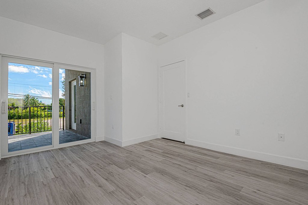 Pearl at Pompano Beach Townhomes in Pompano Beach, FL Bright, empty room with gray wood-look flooring, white walls, sliding glass doors to a balcony with a view of trees and blue sky.