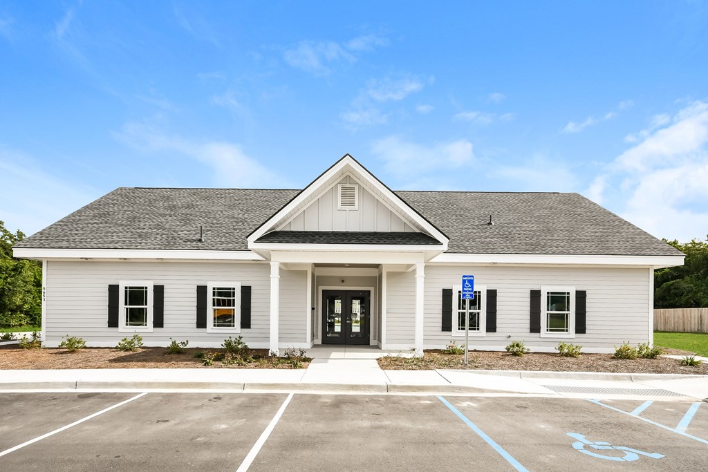 A small white building with a black door and windows, a sign on the door, and a parking lot in front.