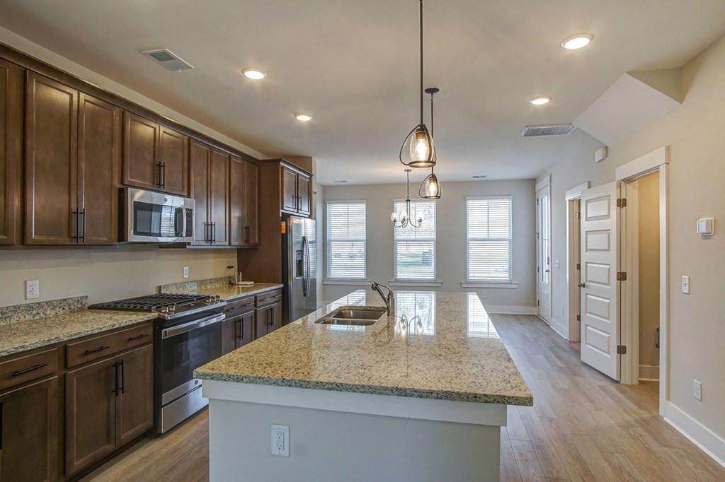 Houses for Rent in North Charleston, SC - Sumner Village - Kitchen with Wood-Style Flooring, Granite Countertops, Stainless Steel Appliances Including a Refrigerator, Oven, and Built-In Microwave, Brown Cabinetry, a Large Kitchen Island, Overhead Hanging Lights, and a View of the Connected Living Area.