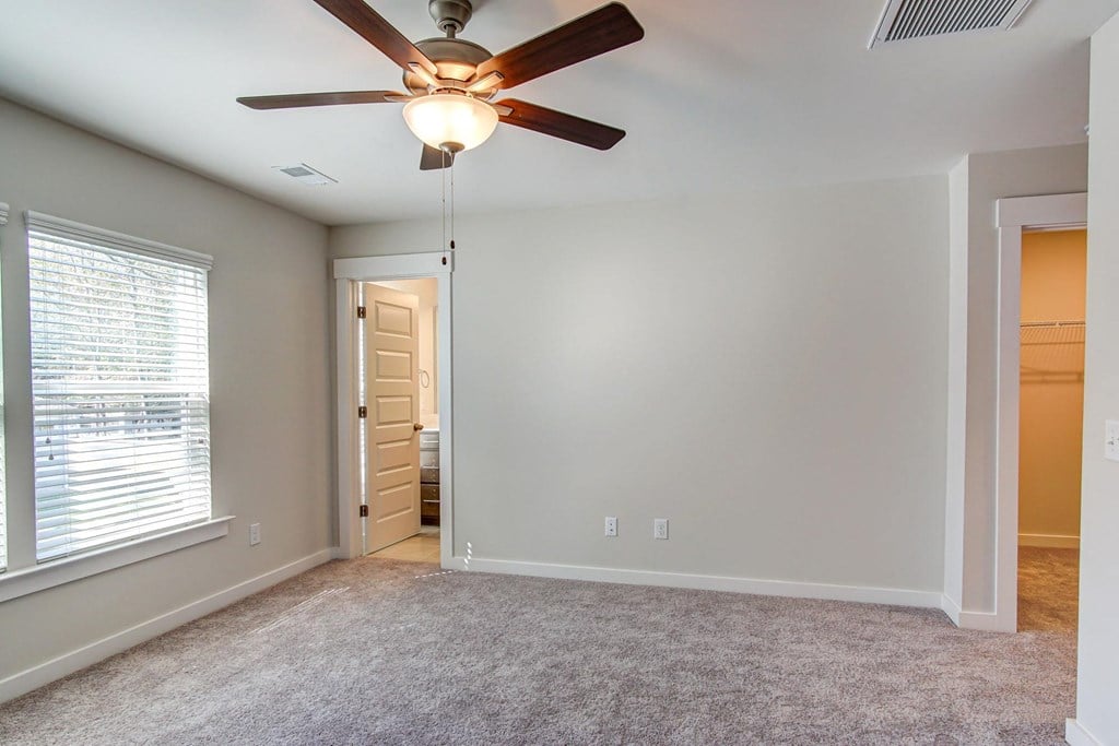 Dog-Friendly Townhomes in Park Circle, SC - Sumner Village - Empty Bedroom with Plush Carpeting, a Ceiling Fan with Lighting, an Attached Bathroom with Tile Flooring, a Spacious Attached Closet, and Large Windows with White Blinds.
