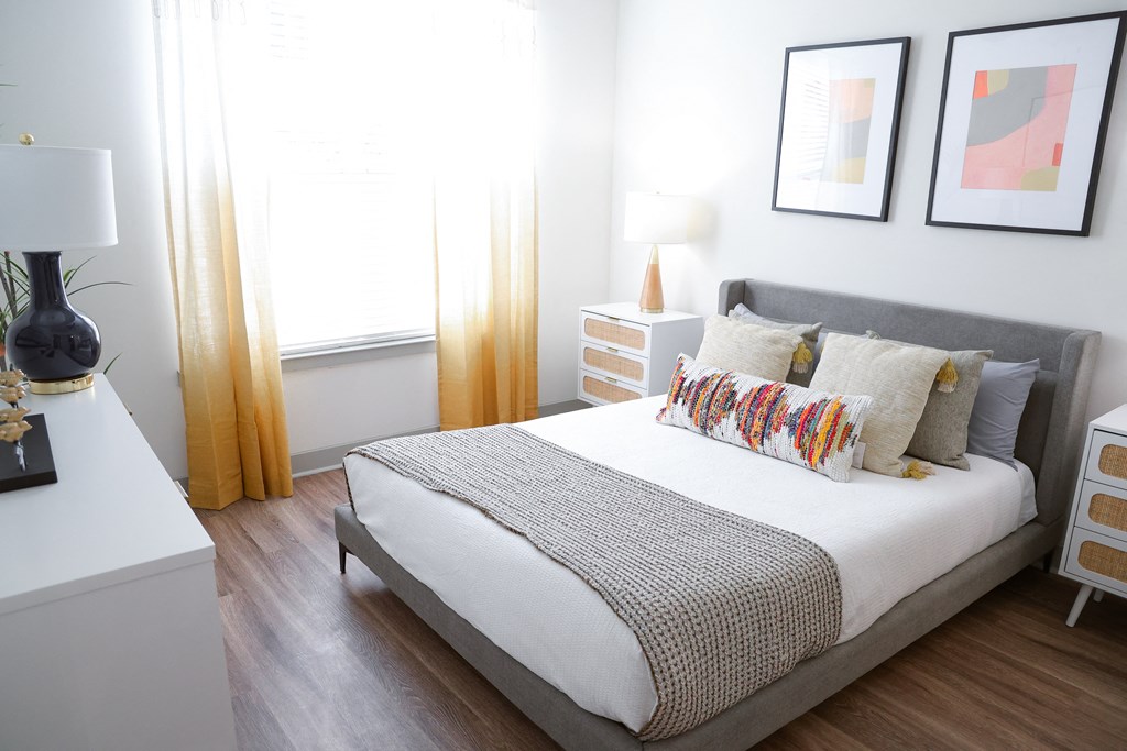 Modern bedroom with gray upholstered bed, white linens, colorful lumbar pillow, and abstract art.