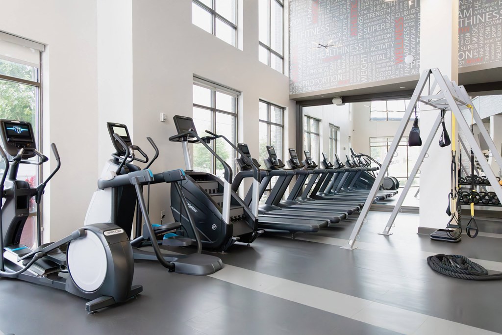 Bright, modern gym featuring treadmills, elliptical machines, a stair climber, and a functional training rig with hanging straps and weights.