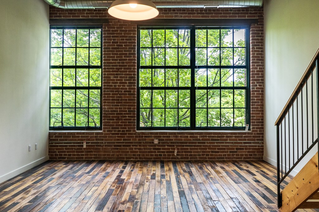 Empty loft apartment with exposed brick, large windows overlooking trees, and hardwood floors.