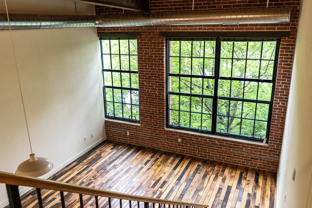 Loft apartment interior with exposed brick, hardwood floors, and large windows overlooking trees.