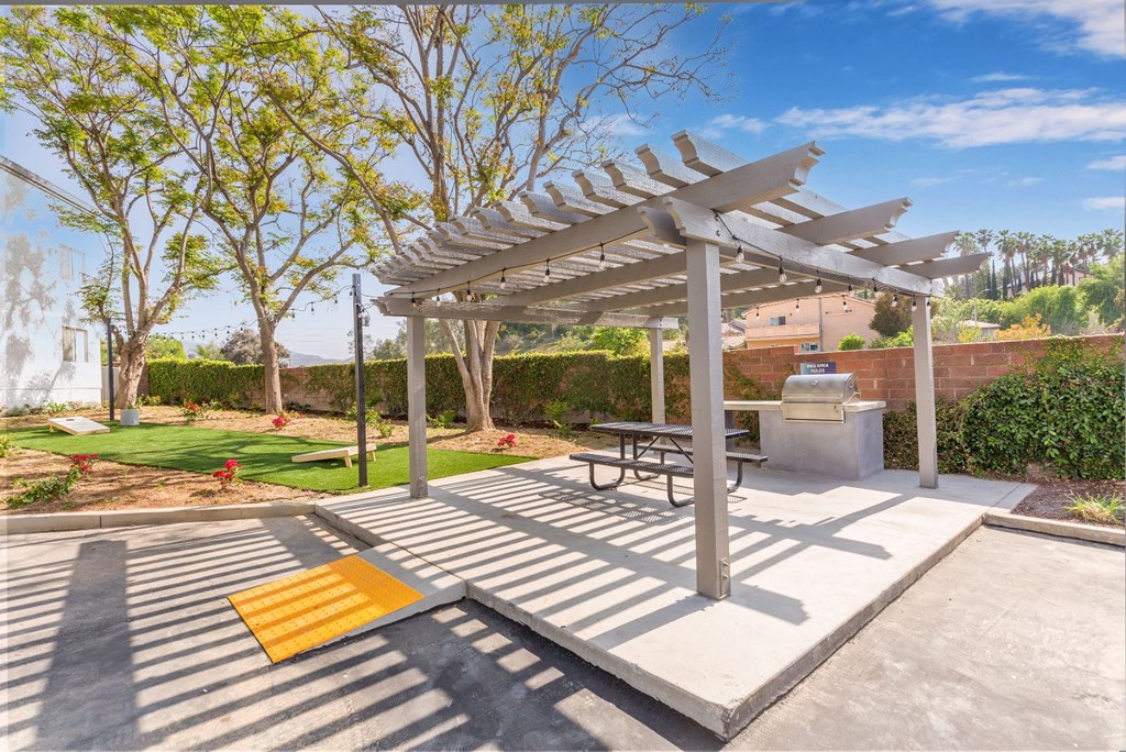 One-Bedroom Apartments In Colton, CA - Ardella At Reche Canyon - Picnic Area Under Pergola With Wheelchair Ramp, BBQ Grill, Table With Benches, Beside Cornhole Area.