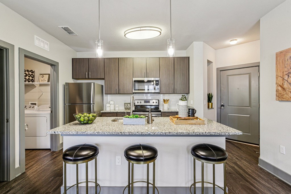 Modern open-concept kitchen with a large island and quartz countertop, featuring three barstools, stainless steel appliances, pendant lighting, and light-toned cabinetry.