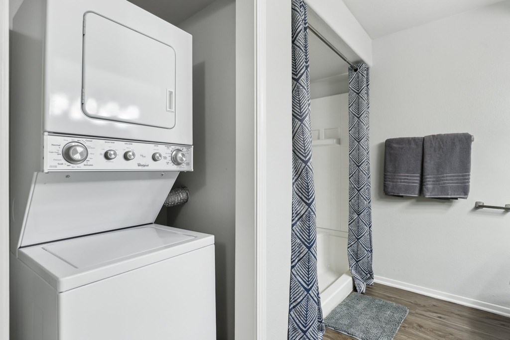 A white washing machine sits next to a bathroom with a grey towel hanging on the wall.