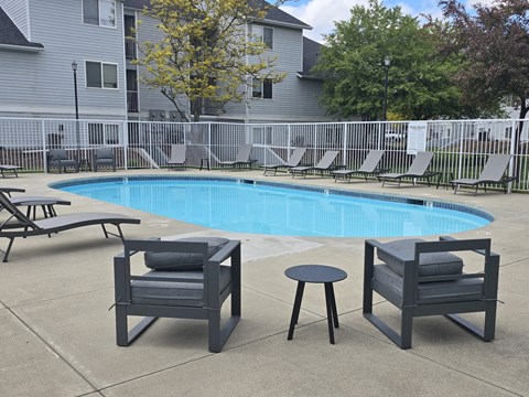 Coeur d'Alene, ID, Apartments for Rent - Trail Lodge Apartments - Outdoor Swimming Pool With Sundeck, Lounge Chairs, Side Table, Patio Chairs, White Safety Fence, and Trees.