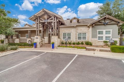 Three Bedroom Apartments in San Antonio TX - Club at Stone Oak - Large Exterior of a Building with Stone Walls, Tan Coloring, Landscaped Plant Beds, and Parking