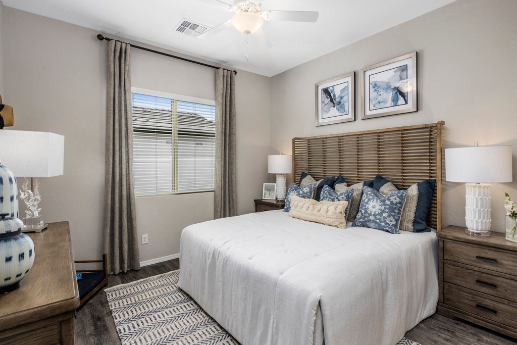 One Bedroom Apartments in Phoenix AZ - The Crofton at Sheely Farms - Furnished Bedroom with a Large Bed, Wooden Nightstand, a Dresser, a Ceiling Fan, Soft Grey Walls, and a Window