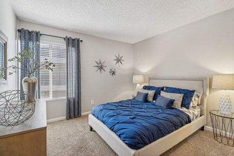 Pet-Friendly Apartments in Downey, CA - Park Regency Club Apartments - A Bedroom With Upholstered Bed, Blue Bedding, Matching Nightstands With Lamps, Carpet Flooring, And A Window.