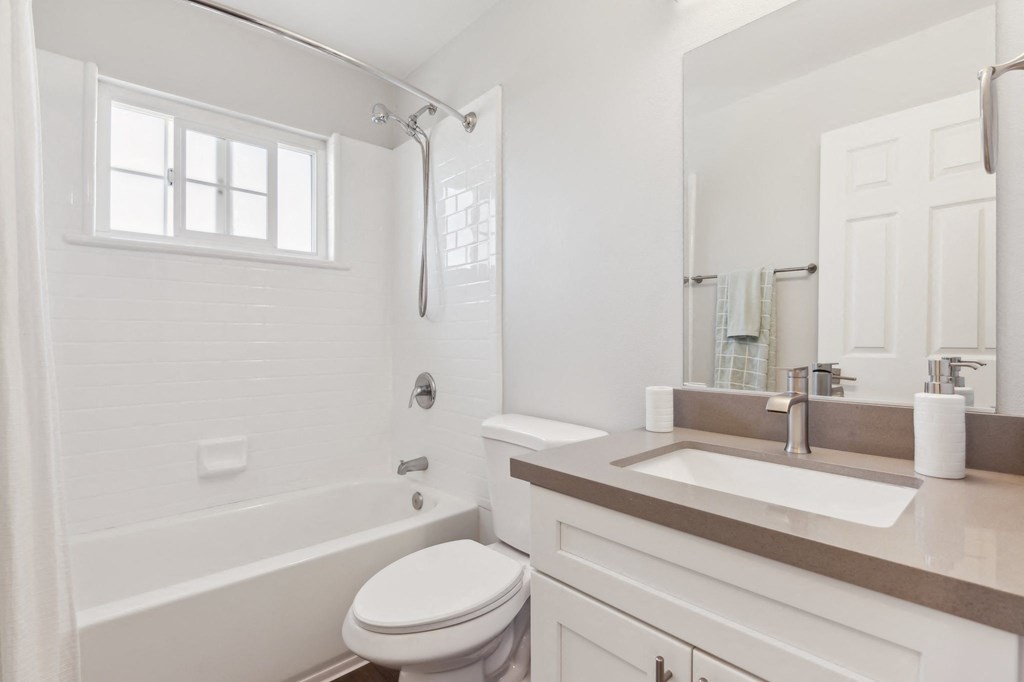 2-Bedroom Apartments in Hayward, CA - Paseo Gardens - Bathroom with Shower and Bathtub Combo with White Tile, A Window, A Sink with White Cabinets, and a Mirror.