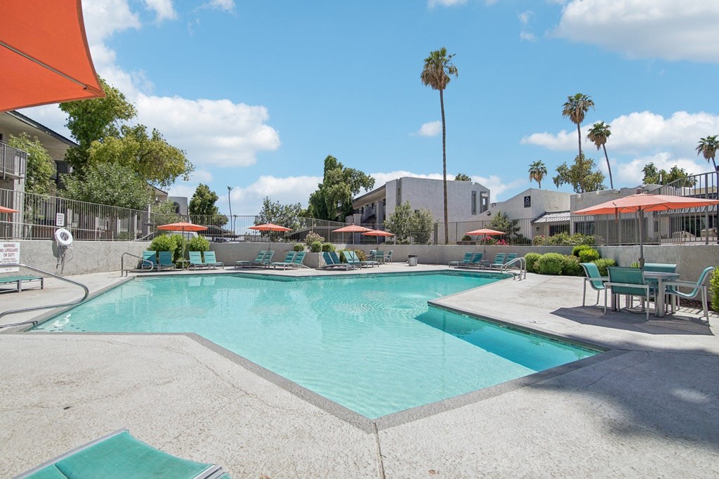 Phoenix Apartments - Ardella on 28th - Gated Outdoor Pool Area with Lounge Seating, Umbrellas, Tables with Chairs, and Surrounding Trees.