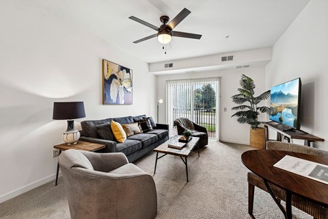 A living room with a grey couch, a grey armchair, a wooden coffee table, a ceiling fan, a flat screen TV, and a painting on the wall.