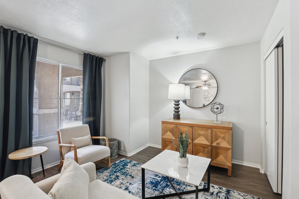A living room with a white sofa, a wooden chair, a white coffee table, a wooden cabinet, a round mirror, and a blue and white rug.