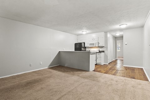 A kitchen with white cabinets and a black fridge is in the background of a living room with a beige carpet.