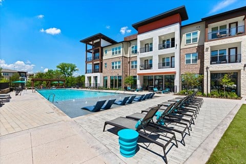 A swimming pool area with sun loungers and apartment buildings in the background.