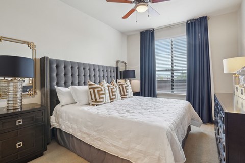 A bedroom with a large bed, a ceiling fan, and a window with blue curtains.