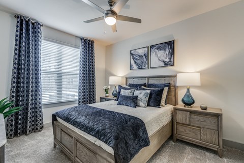 McKinney, TX, Luxury Apartments Near Bucee - Grand at Anna - Primary Bedroom With Plush Bed, Blue Bedding, Nightstands With Lamps, Ceiling Fan, and Large Window With Patterned Curtains.