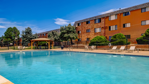 A large swimming pool in front of a brown building with a fence and trees.