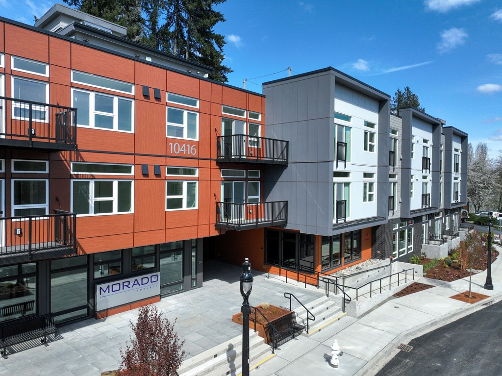 Studio Apartments in Bothell, WA - Morado Bothell - Street View of Property Exterior with Red and Grey Walls, Balconies, Sidewalk, and Street Lamps.
