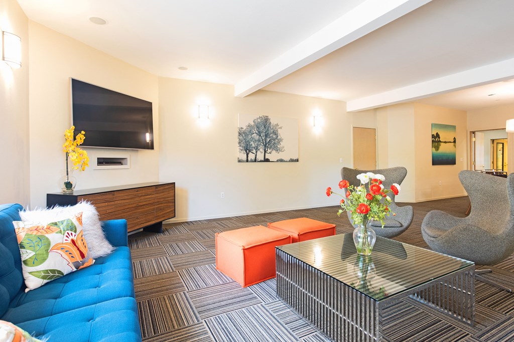 Puyallup WA Apartments - One Canyon Place - Clubhouse with  Wide-Screen TV, Plush Couch, Two Square Stools, Two Wingback Chairs, and Glass-Topped Coffee Table