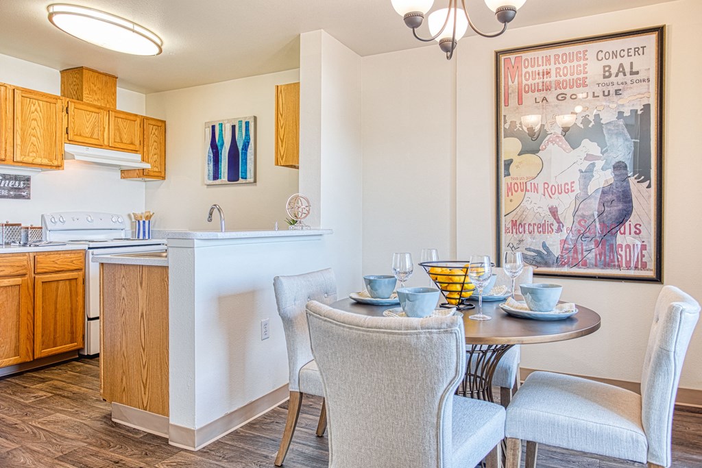Apartments in Puyallup, WA, for Rent - One Canyon Place - Dining Area with Wood-Style Flooring, Round 4-Seater Dining Table, and View of Kitchen with White Electric Range, High Bar Countertop, and Wooden Cabinets.