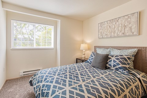 Three-Bedroom Apartments in Puyallup, WA - One Canyon Place - Comfortable Bedroom With Large Window and Blinds, Plush Carpet, Nightstand, Lamp, and Patterned Bedding With Accent Pillows.