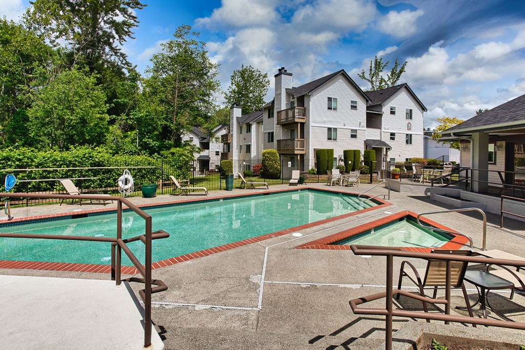Garden-Style Apartments in University Place, WA, with In-Unit Washer and Dryer - Carriage House - Swimming Pool Adjacent to a Hot Tub, Surrounded by Lounge Chairs, Trees, and Apartments.