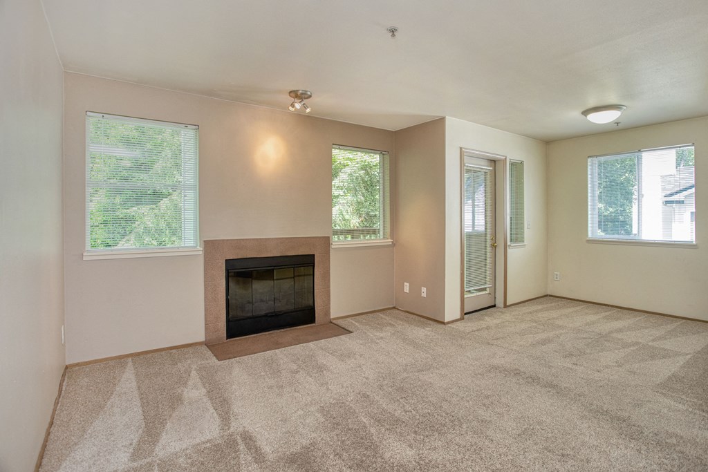 Pet-Friendly Garden-Style Apartments in University Place, WA - Carriage House - An Empty Living Room with Carpet Flooring, Wood Burning Fireplace, Light Fixtures, Windows, and a Glass-Paneled Door with Blinds.