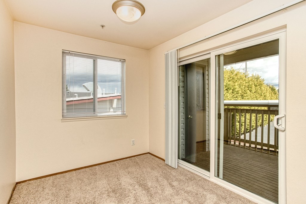 University Place, WA, Garden-Style Apartments - Carriage House - An Empty Room with Carpet Flooring, Light Fixture, Window, and Sliding Glass Door with Blinds, Leading to a Balcony.