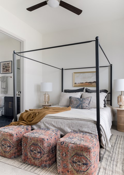 2-Bedroom Luxury Apartments in Uptown, Dallas, TX - Katy Trail Uptown - Bedroom With Black Canopy Bed, Layered Bedding, Patterned Ottomans, Side Tables, And Attached Bathroom.