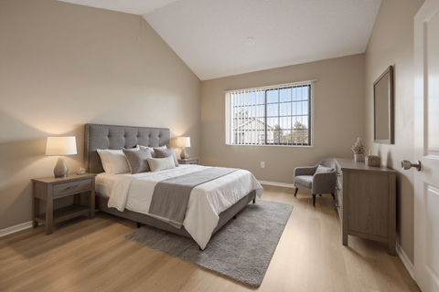 Aurora, CO, Apartments Near Denver Zoo - The Juliet at Lowry - Bedroom with Plank Flooring, Bed, Nightstands, Lamps, Dresser, Armchair, Wall Frame, and a Large Window with Blinds.