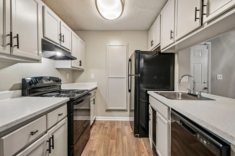 A black refrigerator stands in a kitchen with white cabinets and a wooden floor.