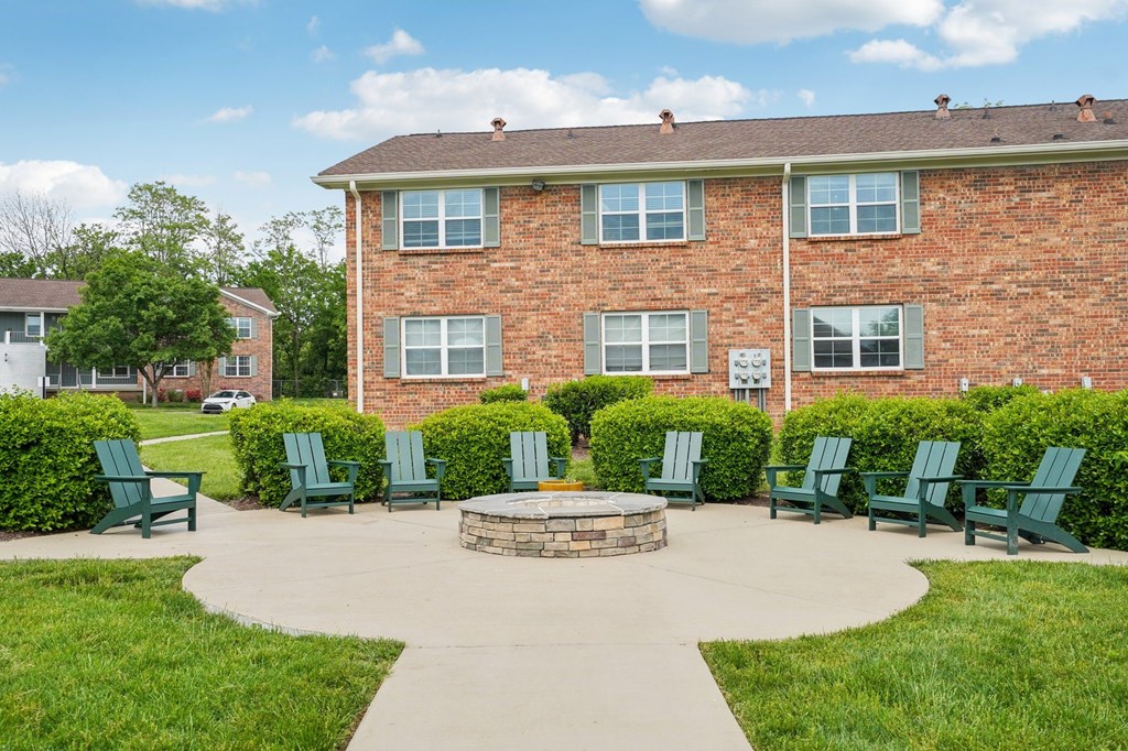 Outdoor patio with stone fire pit and green Adirondack chairs in front of a brick apartment building.