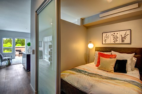 Bothell Apartments - 98th Avenue - A Bedroom With A Sliding Frosted Glass Door, Layered Pillows, Floor Lamp, And Wood-Style Flooring.
