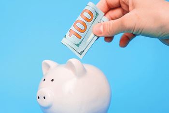 A hand is putting a 100 dollar bill into a piggy bank.