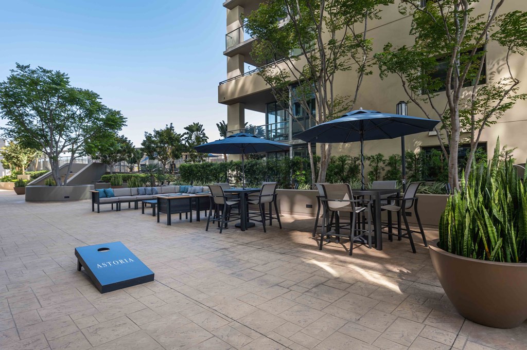 Apartments for Rent in Irvine, CA - Astoria at Central Park West - Outdoor Courtyard with Lounge Seating, Umbrella Tables, Chairs, Cornhole Board, Potted Plants, Trees.