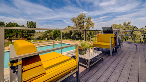 Aurora, CO apartments - Aurora Hills - a poolside deck with yellow chairs, a table, and a railing overlooking a blue pool and various types of trees.