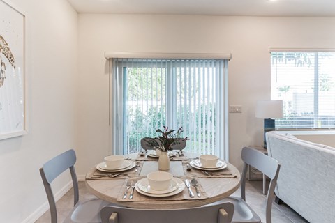 Dining room with round table set for four, light gray chairs, and vertical blinds.
