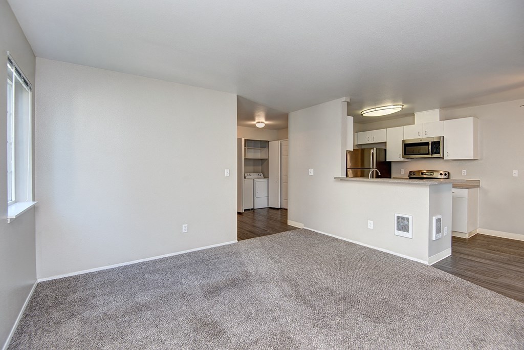 the living room and kitchen of an apartment with white walls and carpet
