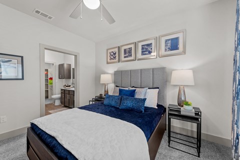 Cibolo, TX, Apartments Near Retama Park - Grand at Cibolo - Cozy Bedroom With Upholstered Headboard, Blue Bedding, Bedside Lamps, And Ensuite Bathroom Entryway.
